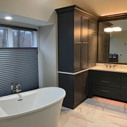 A master bathroom with a freestanding bathtub and a double sink with a large mirror above them. There are several cabinets to the left of the sinks and drawers underneath. The floors are a white and grey marbled porcelain and the walls are off-white.