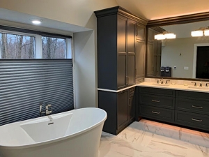 A master bathroom with a freestanding bathtub and a double sink with a large mirror above them. There are several cabinets to the left of the sinks and drawers underneath. The floors are a white and grey marbled porcelain and the walls are off-white.