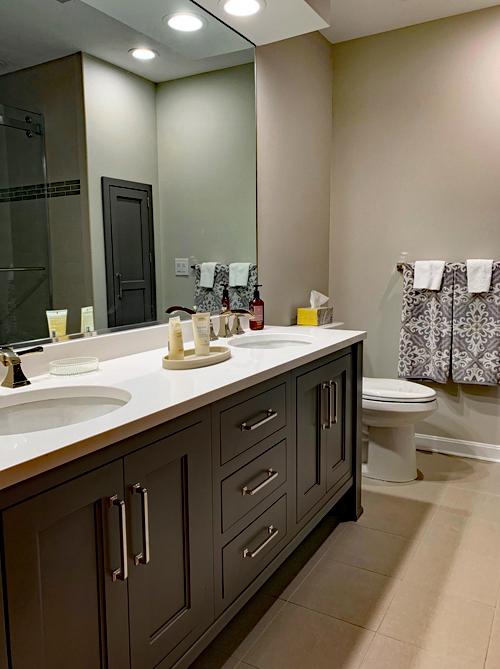 Another bathroom in the same house with double sinks. There is a commode to the right and a towel rack on the wall. In the mirror you can see the corner of a walk-in shower with glass doors. The floors are an off-white tile and the walls are painted a similar off-white color.