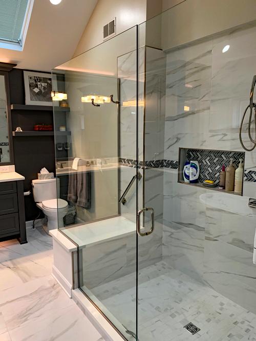 A large walk in shower with glass doors. The shower walls and floor are the same marbled porcelain tiling as the floors of the rest of the bathroom. There is a built in caddy to hold soap. There is also a seated area in the shower with a grab bar.