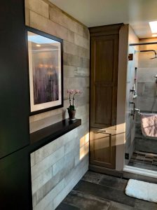 A master bathroom with a highlight wall next to the walk in shower. The wall is a beautiful textured light grey tile to contrast the dark grey textured tile floor. There is a shelf built in to the wall. A potted plant sits on the shelf and a large framed painting hangs above it. There is a narrow space of wall between this and the shower where John has installed a cabinet and drawer.