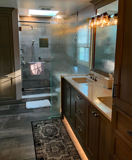 The right side of the same bathroom. There is a double sink with plenty of cabinet and drawer space underneath. Behind the sink is a fogged-glass partition which hides the commode. Behind that is the large walk-in shower that includes both a fixed and removable shower head and a grab bar. There is a skylight on the ceiling of the shower and the rightmost wall in the shower has a fogged glass window that let natural light into the room.